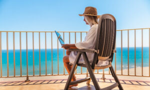 Woman working on a laptop on a balcony overlooking a beach.